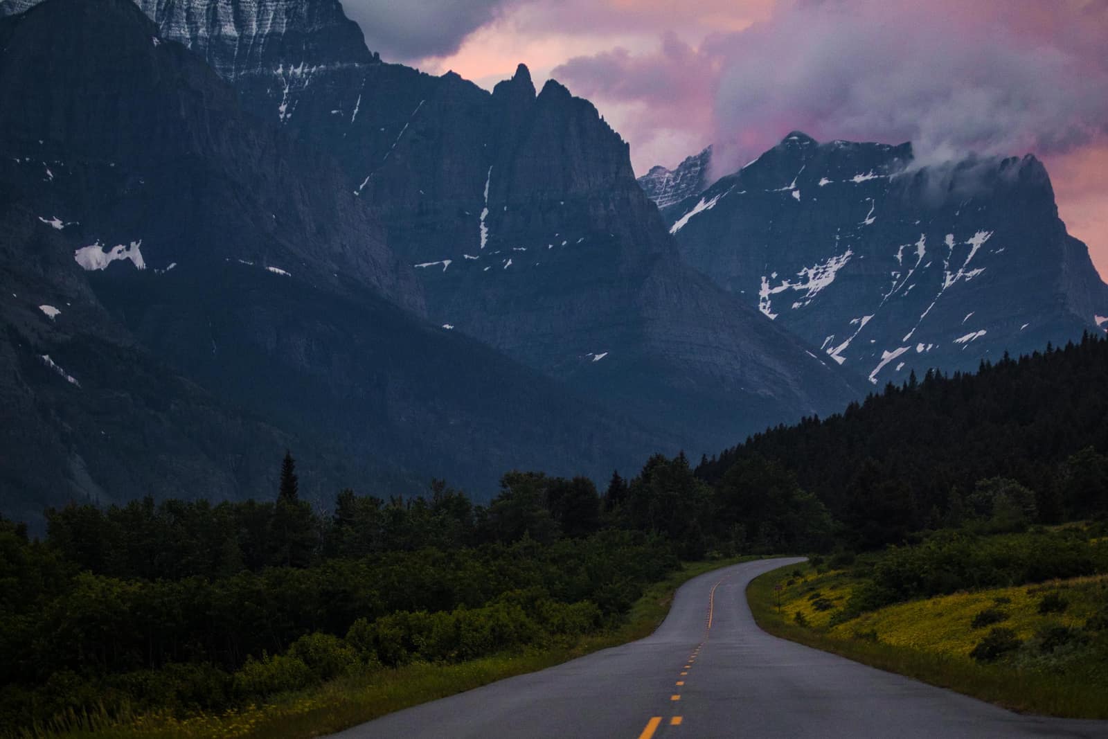 A two-lane road curves toward the jagged peaks of Glacier National Park at sunset, with snow-patched mountains rising above the forested valley
