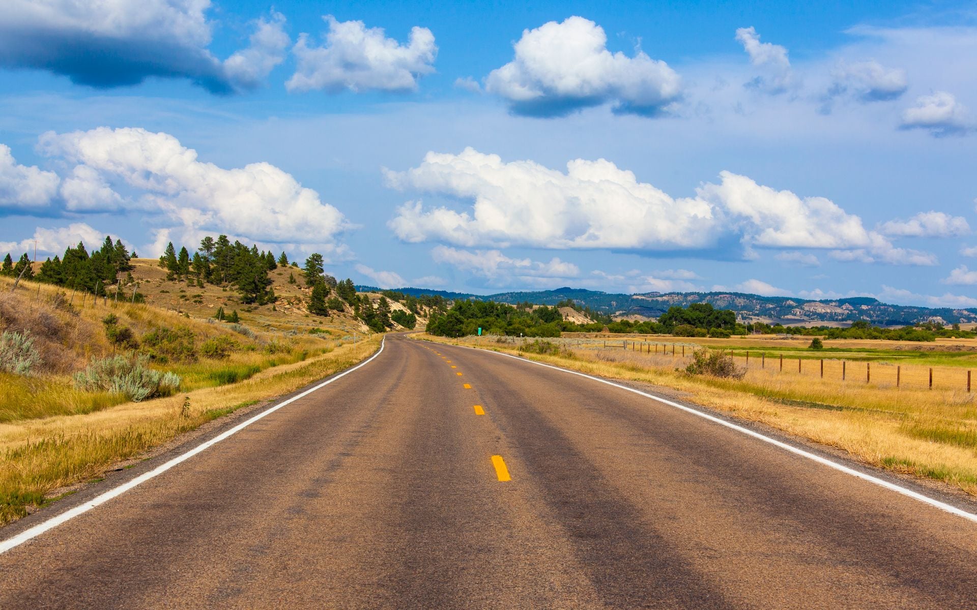 A two-lane highway stretching toward the Rocky Mountain Front under a wide Montana sky