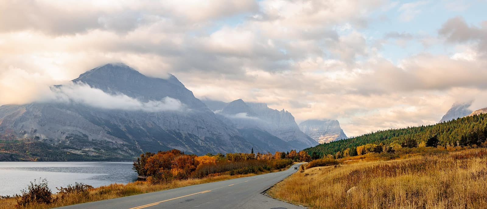A road curving alongside St. Mary Lake in fall, with layered peaks rising through low clouds in Glacier National Park, Montana