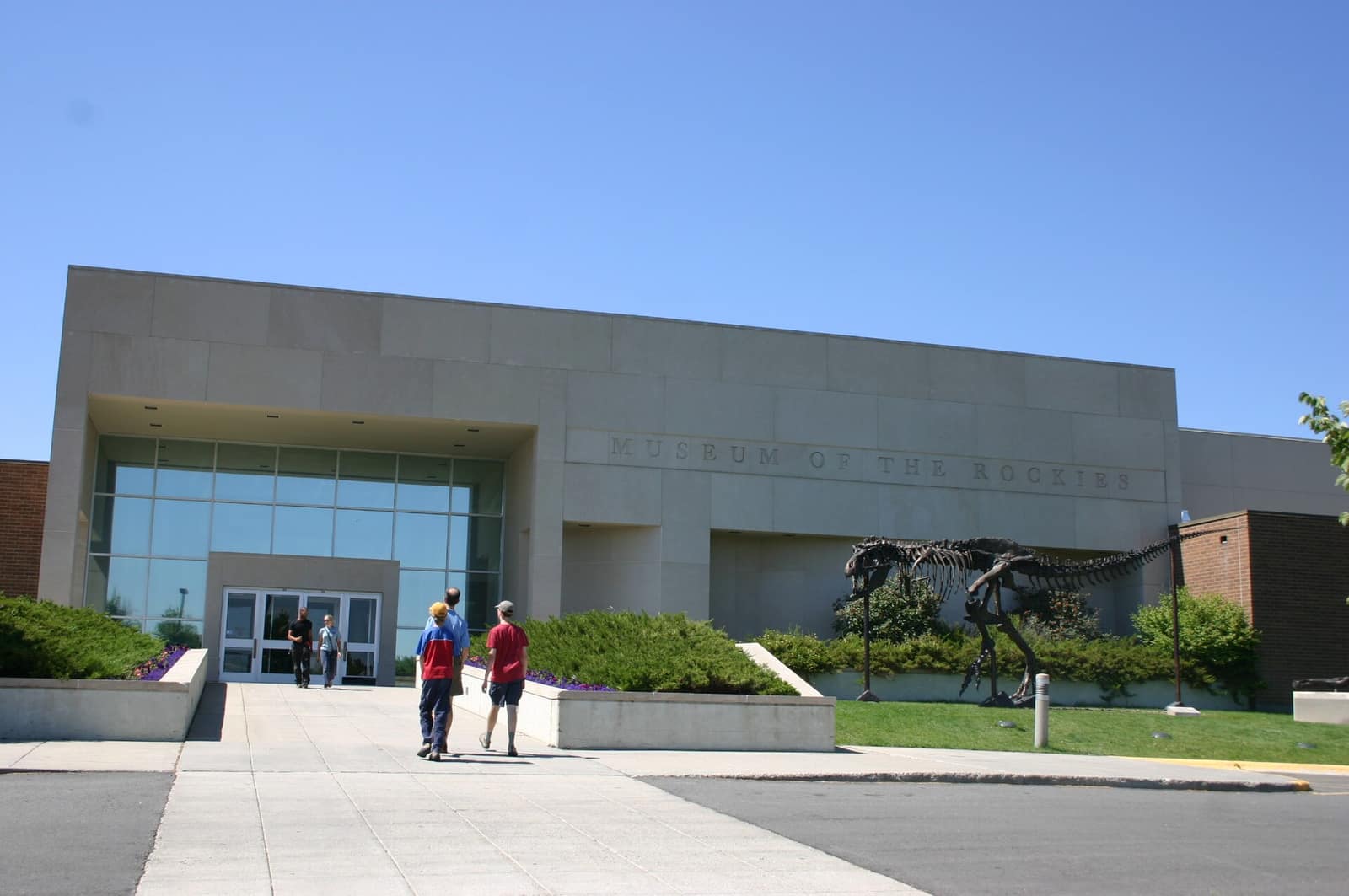 Exterior of the Museum of the Rockies in Bozeman, Montana, with a dinosaur skeleton on the lawn and visitors walking toward the entrance